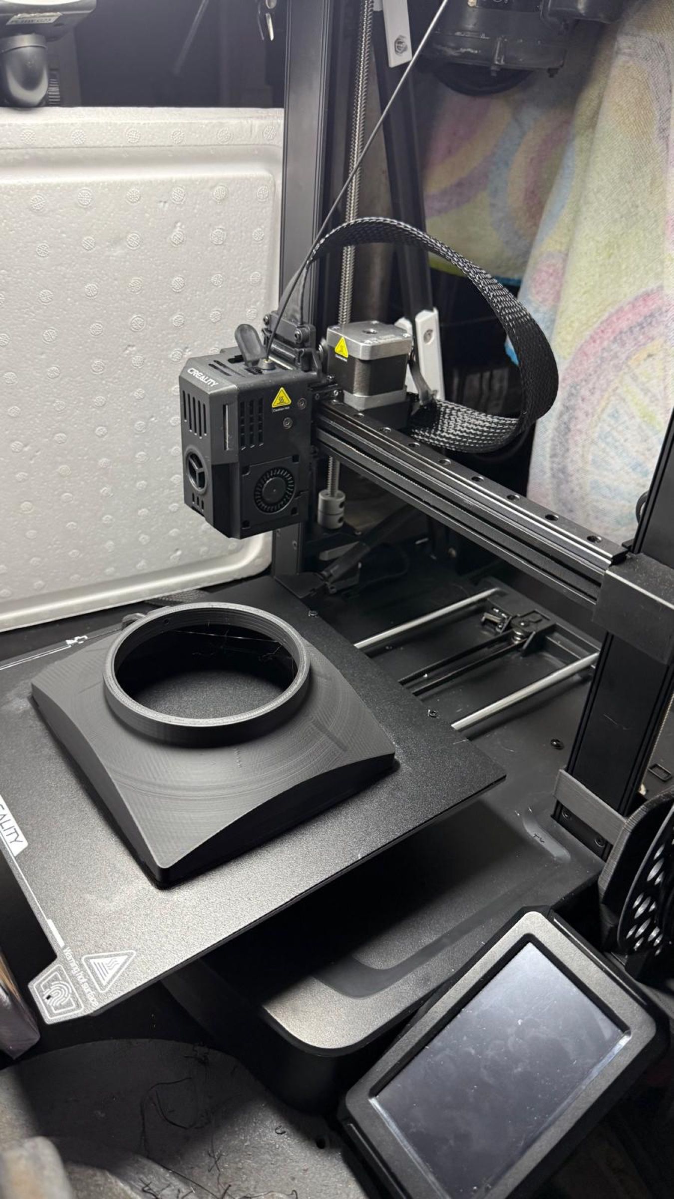 Pieza negra con forma de parasol rectangular y un aro circular, sobre la base de una impresora 3D.