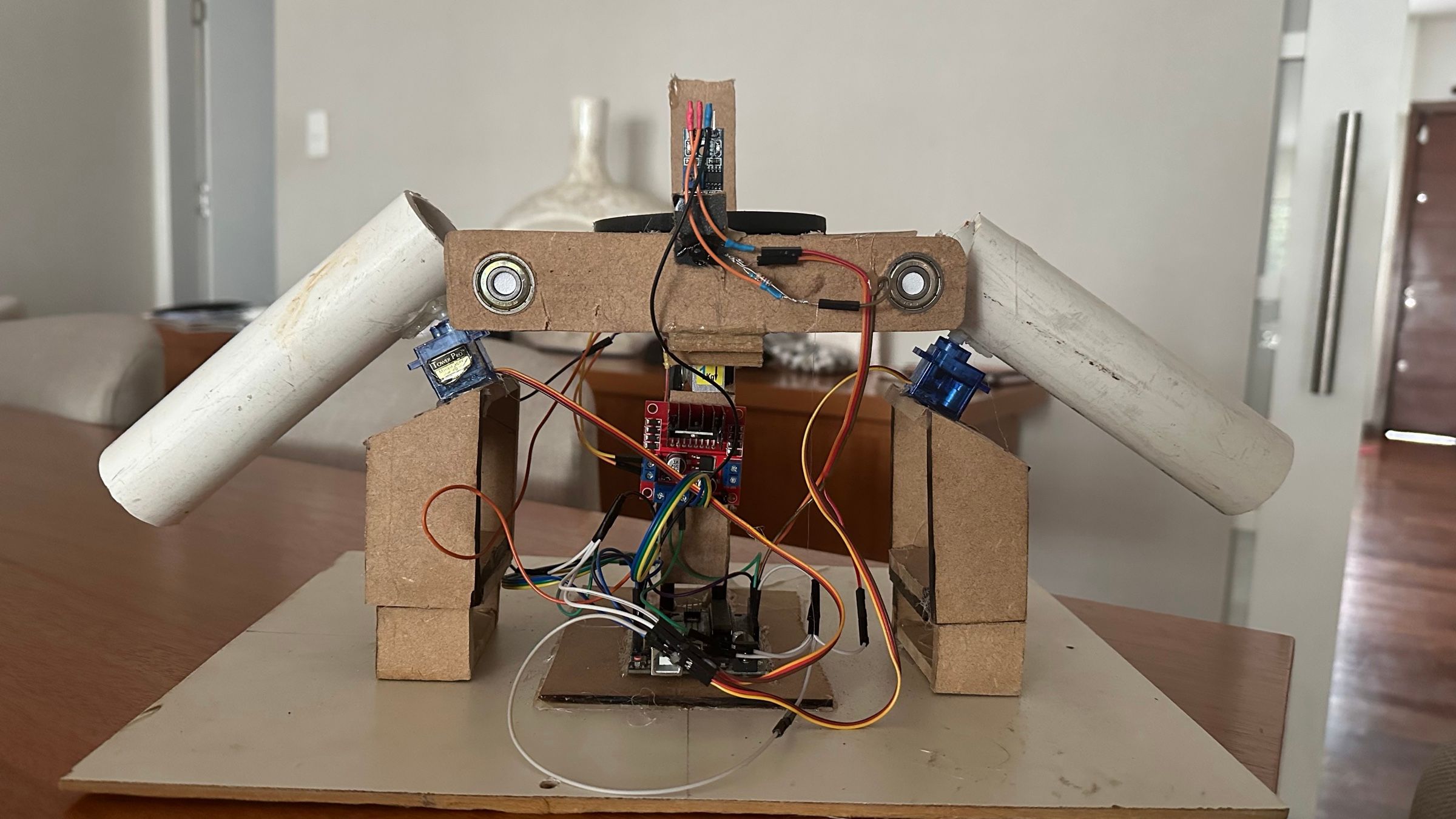 Montaje de cartón y tubos blancos con dos servomotores azules, una placa de control y cables, visto de frente sobre una mesa.