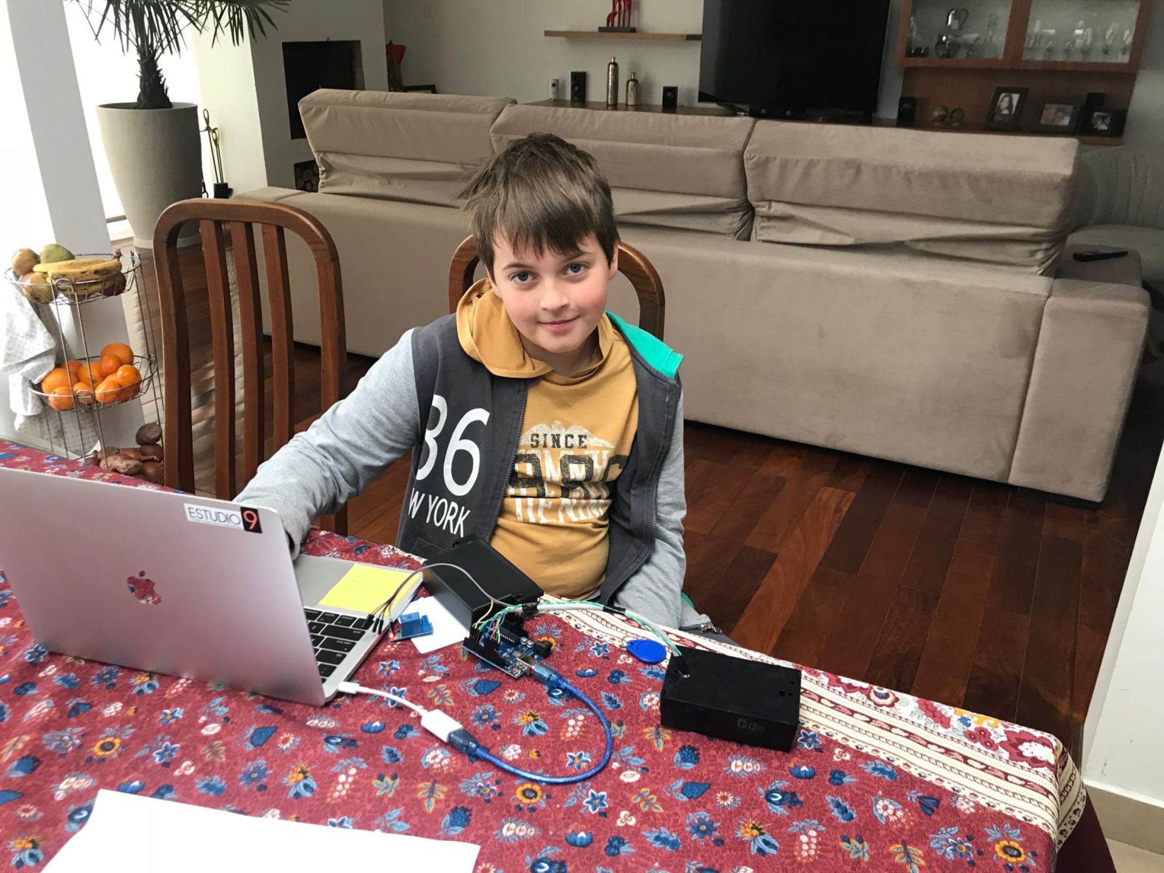 Niño sonriente sentado a una mesa, junto a una laptop, una placa Arduino, un módulo de relé, cables y una etiqueta redonda azul.