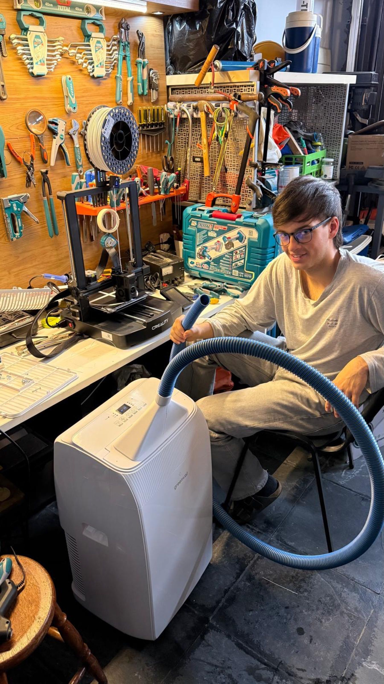 Persona sentada en un taller junto a un equipo de refrigeración portátil con una manguera azul; detrás, una impresora 3D y herramientas colgadas.