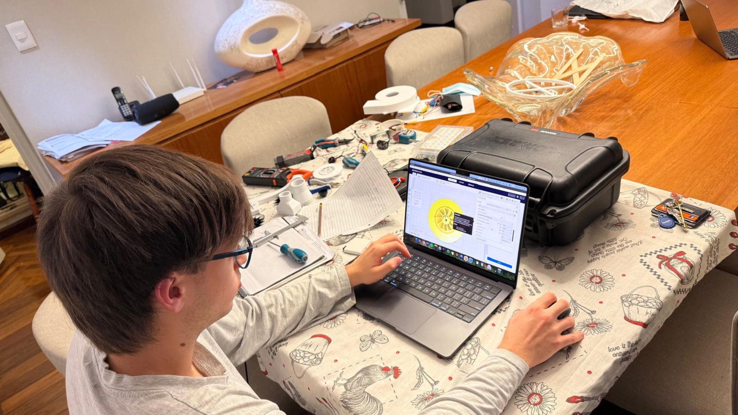Persona con anteojos trabajando en una laptop con un diseño en pantalla, en una mesa con herramientas y componentes electrónicos.